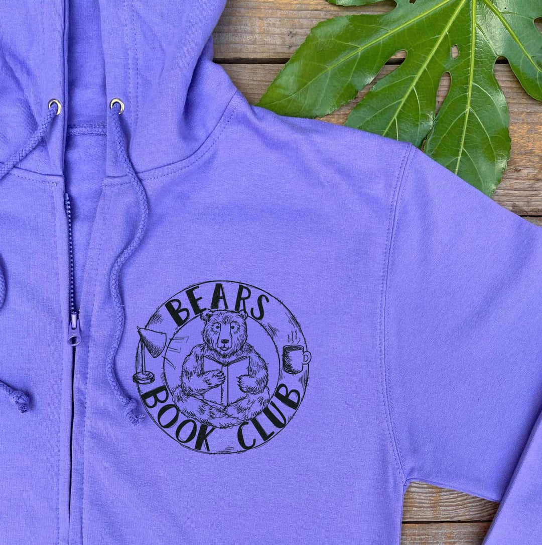 A lavender-purple zip-up hoodie laid flat on a rustic wooden surface, featuring a black circular graphic on the left chest that reads &