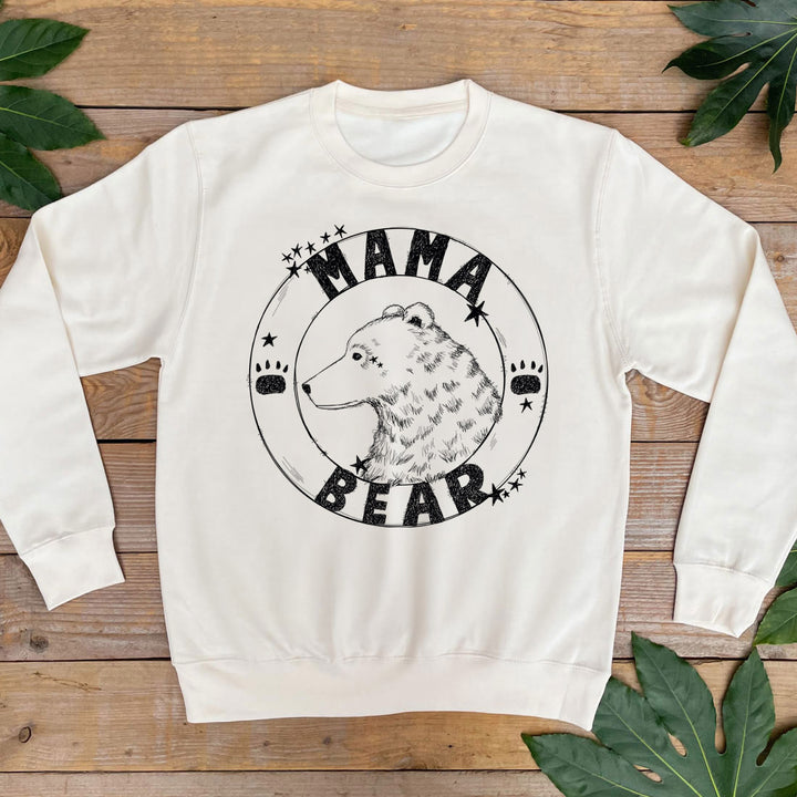 White sweatshirt with 'Mama Bear' graphic on a wooden surface with leaves.